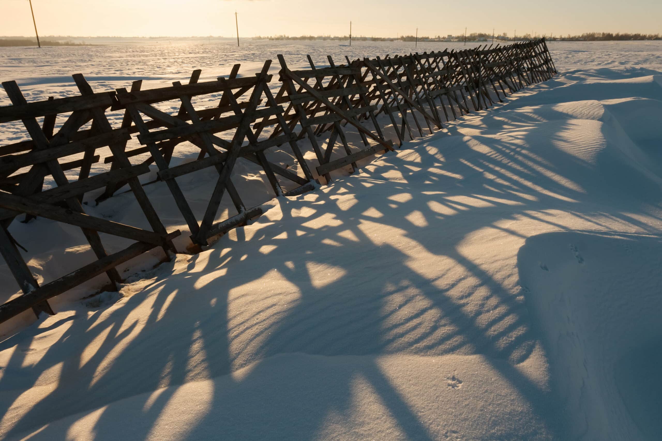 A wooden fence casts long, crisscrossing shadows over rippled snowdrifts in a sunlit, open winter landscape—an inviting view for those seeking hunting property or a ranch for sale under a wide, clear sky.