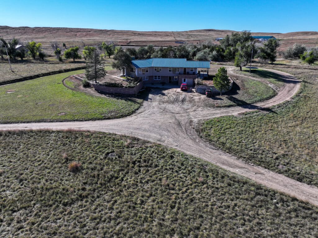 Aerial view of a ranch-style house with a circular driveway, set on open land ideal as a hunting property or cattle ranch. Surrounded by grassy fields and trees under a clear blue sky, two vehicles are parked nearby.