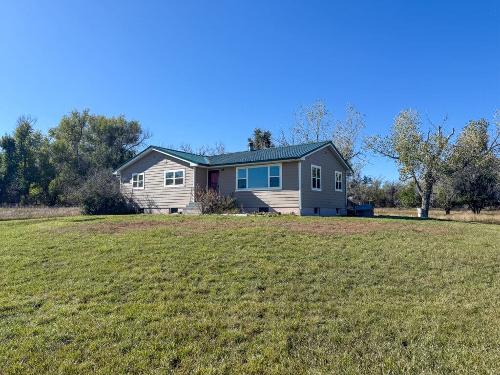 A single-story beige house with a green roof sits on a grassy lawn under a bright blue sky, surrounded by trees and open land—perfect for recreational land or as a hunting property.
