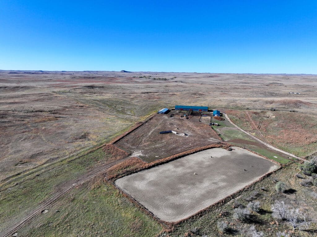Aerial view of a rural cattle ranch with several buildings, fenced areas, and a large rectangular riding arena on open, flat grassland under a clear blue sky. Ideal recreational land for those seeking a classic ranch for sale.