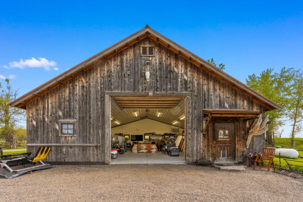 A rustic wooden barn with an open central door reveals tools and equipment inside. Set on gravel, the barn sits amid green trees under a clear blue sky—ideal recreational land or hunting property, with outdoor chairs by the entrance.