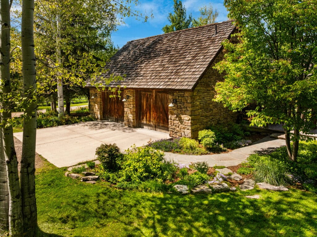 A rustic stone garage with three wooden doors sits among lush green trees on a picturesque cattle ranch, with sunlight casting shadows across the landscaped gardens, driveway, and lawn.