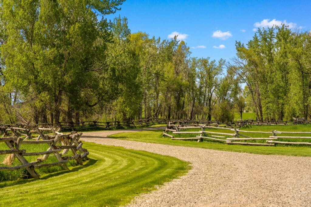 A winding gravel path curves through lush green grass, bordered by wooden fences and tall leafy trees under a bright blue sky—a picturesque ranch for sale perfect for those seeking beautiful recreational land.