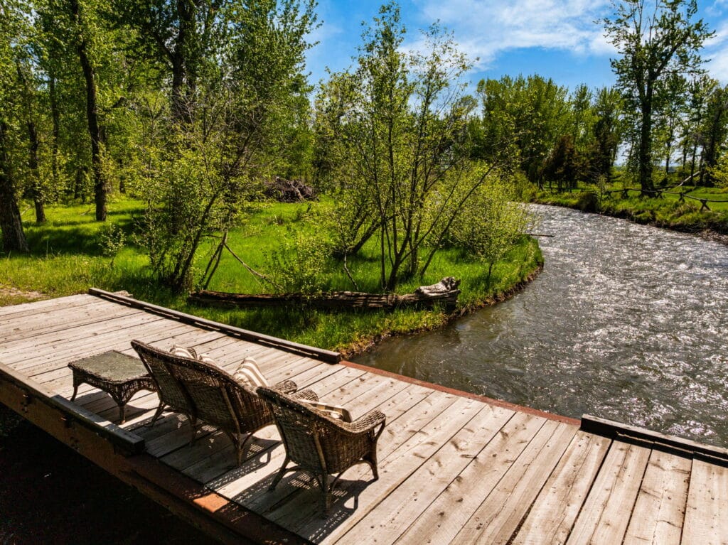 Two wicker chairs and a table sit on a wooden deck by a winding river, surrounded by green trees and grass under a bright blue sky—perfect for relaxing on your dream hunting property or cattle ranch.
