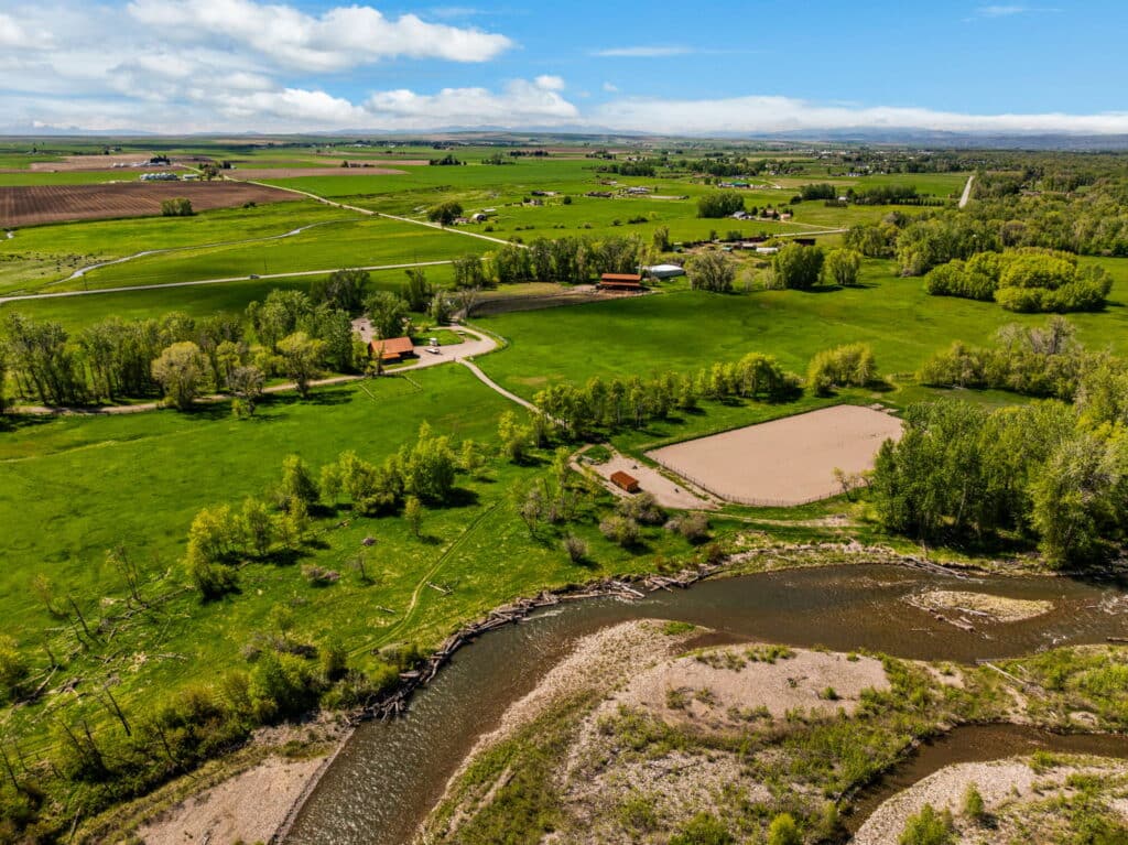 Aerial view of green fields, scattered trees, farm buildings, and a winding river with a sandy bank under a partly cloudy sky in a rural landscape—ideal hunting property or ranch for sale.
