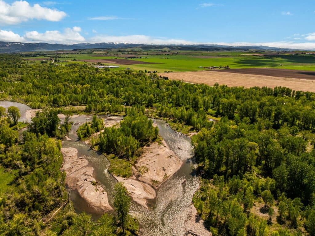 Aerial view of a winding river flowing through lush green forest and open fields with distant mountains under a blue sky—a prime example of recreational land or ranch for sale.