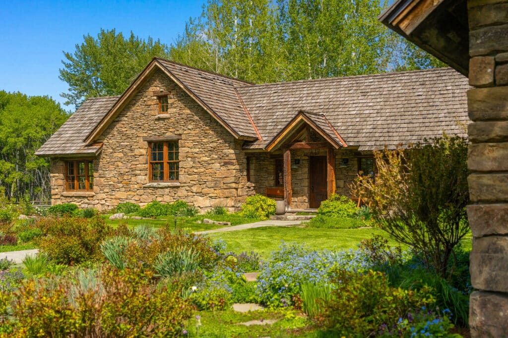 A rustic stone cottage with a wooden roof sits surrounded by lush greenery, colorful flowers, and a well-maintained garden under a bright blue sky—an inviting retreat perfect for recreational land enthusiasts.