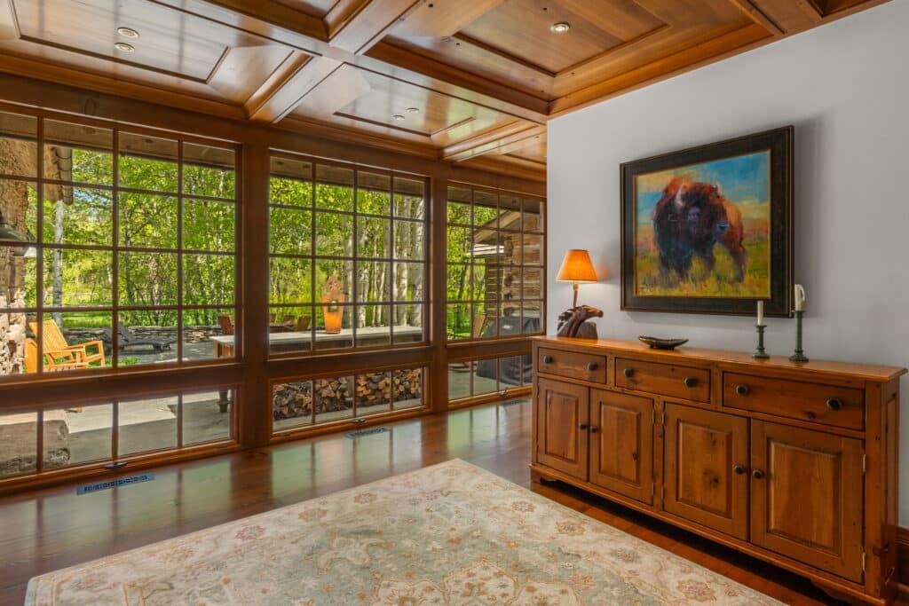 Bright room with large floor-to-ceiling windows, wooden ceiling and floors, a rustic sideboard with lamp, candles, and bison painting above. Patio with chairs and greenery outside. Ideal setting for a hunting property or cattle ranch for sale.