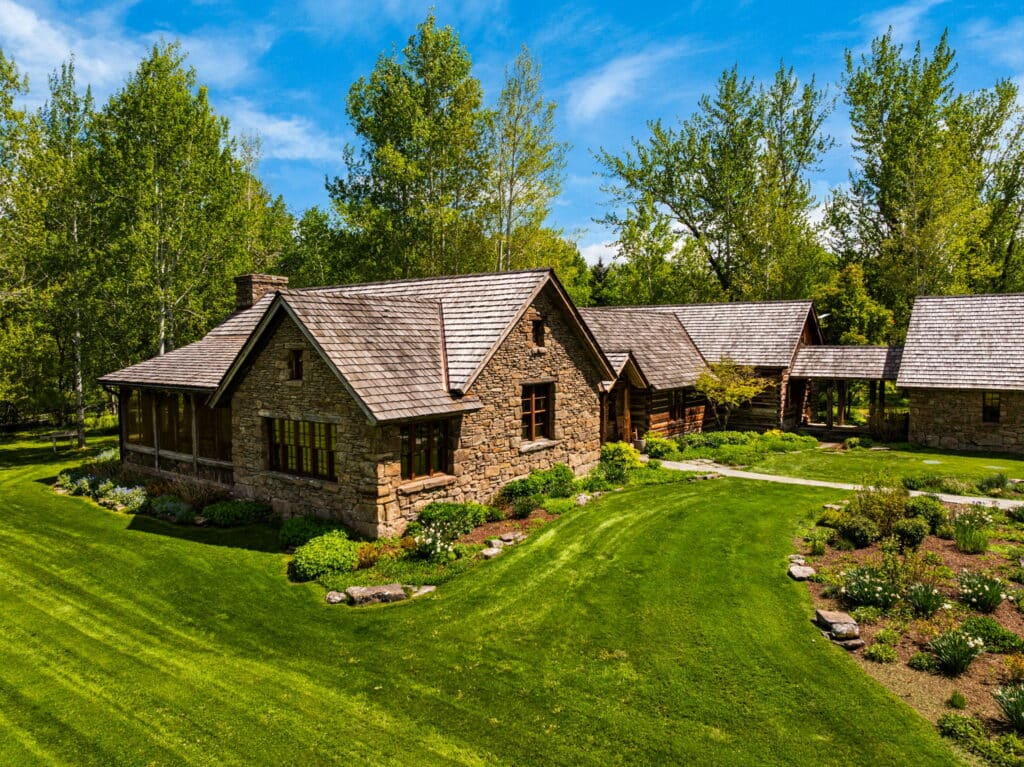 A rustic stone and wood cabin on a scenic cattle ranch with a well-kept lawn, flower beds, trees, and a blue sky with scattered clouds. A covered walkway connects the main house to another building. Land for sale.