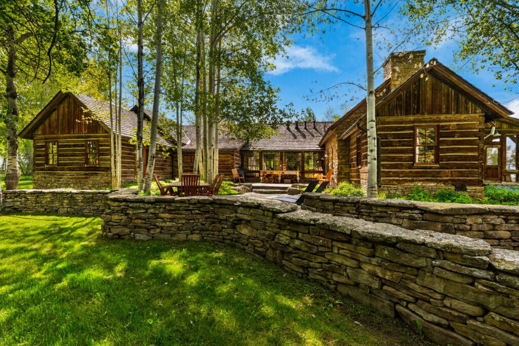 A rustic log cabin with a stone chimney sits among tall trees and green grass, part of a scenic ranch for sale. A stone wall and wooden outdoor chairs rest in the foreground under a bright blue sky.