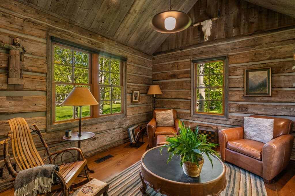 Cozy rustic living room with wood-paneled walls, two leather armchairs, a round coffee table with a plant, a rocking chair, lamps, and large windows letting in natural light—perfect for relaxing after exploring your new hunting property.