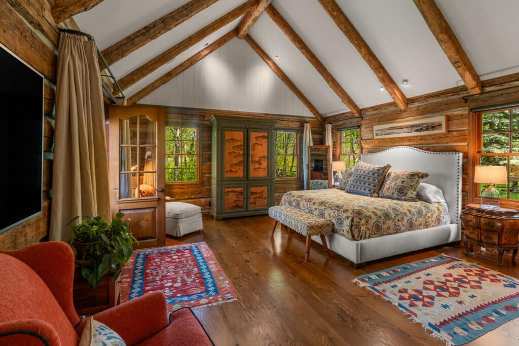 A cozy rustic bedroom with exposed wood beams, a large bed with patterned bedding, antique furniture, colorful rugs, and wood floors. Large windows overlook green trees, perfect for relaxing after a day on a cattle ranch or recreational land. Warm lighting adds charm.