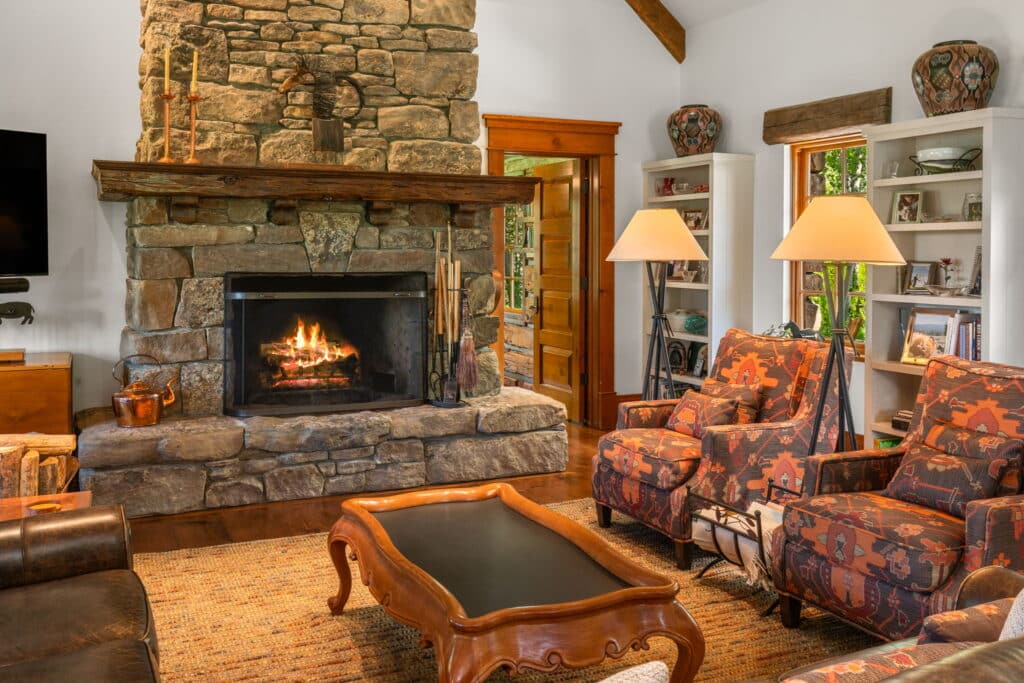 Cozy living room with a stone fireplace, two patterned armchairs, a wooden coffee table, and built-in shelves. Perfect for relaxing after a day on your cattle ranch or exploring your hunting property, with warm lighting from large lamps.