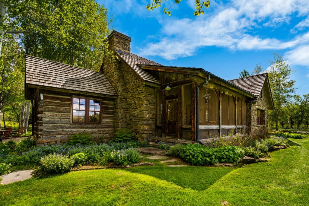 A rustic wooden cabin with a stone chimney sits surrounded by green grass, bushes, and tall trees under a clear blue sky. Sunlight casts shadows across the lush yard of this cozy hunting property and tranquil recreational land.