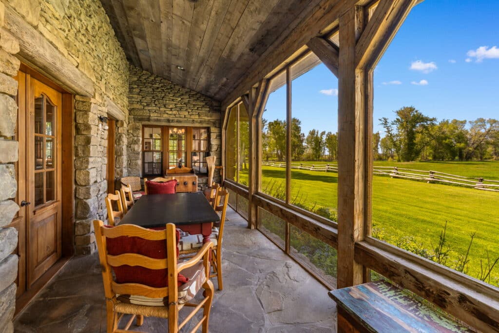 A rustic screened porch with stone walls and wooden beams offers a dining area with views of the lawn and trees under a bright blue sky—ideal for enjoying life on a picturesque cattle ranch or hunting property.