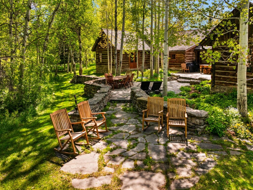 Wooden chairs are arranged on a stone patio surrounded by lush green trees and grass, with rustic log cabins in the background—an inviting ranch for sale on a sunny day.