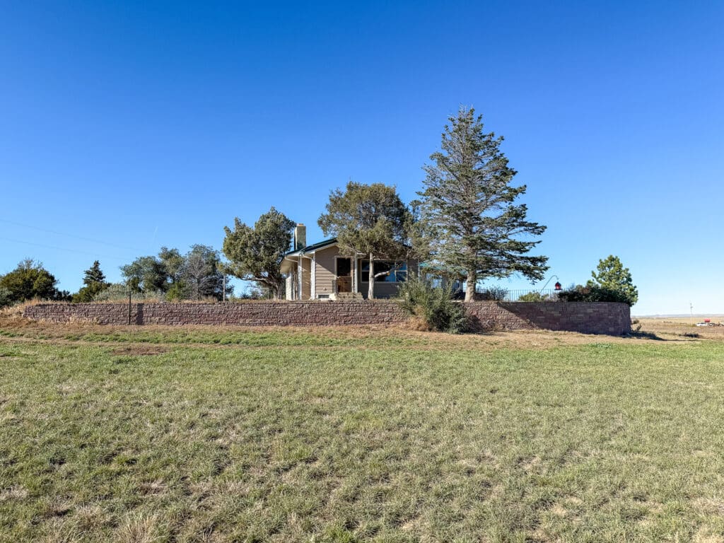 A small ranch for sale sits on a raised stone platform surrounded by trees, with a grassy field in the foreground under a clear blue sky.