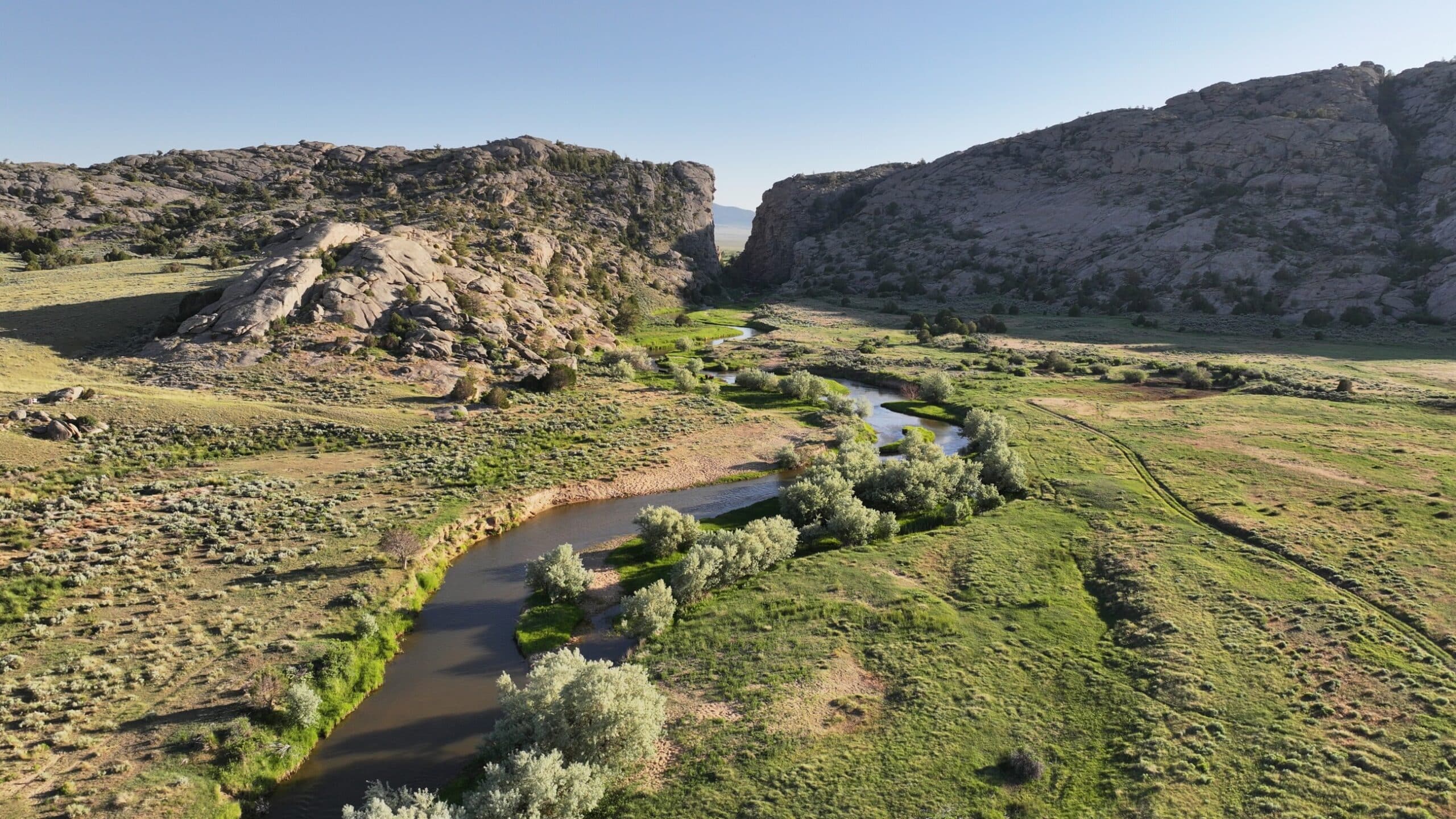A winding river flows through a green valley flanked by rocky hills under a clear blue sky. Sparse trees and shrubs dot this recreational land, and sunlight bathes the scene in a warm glow.