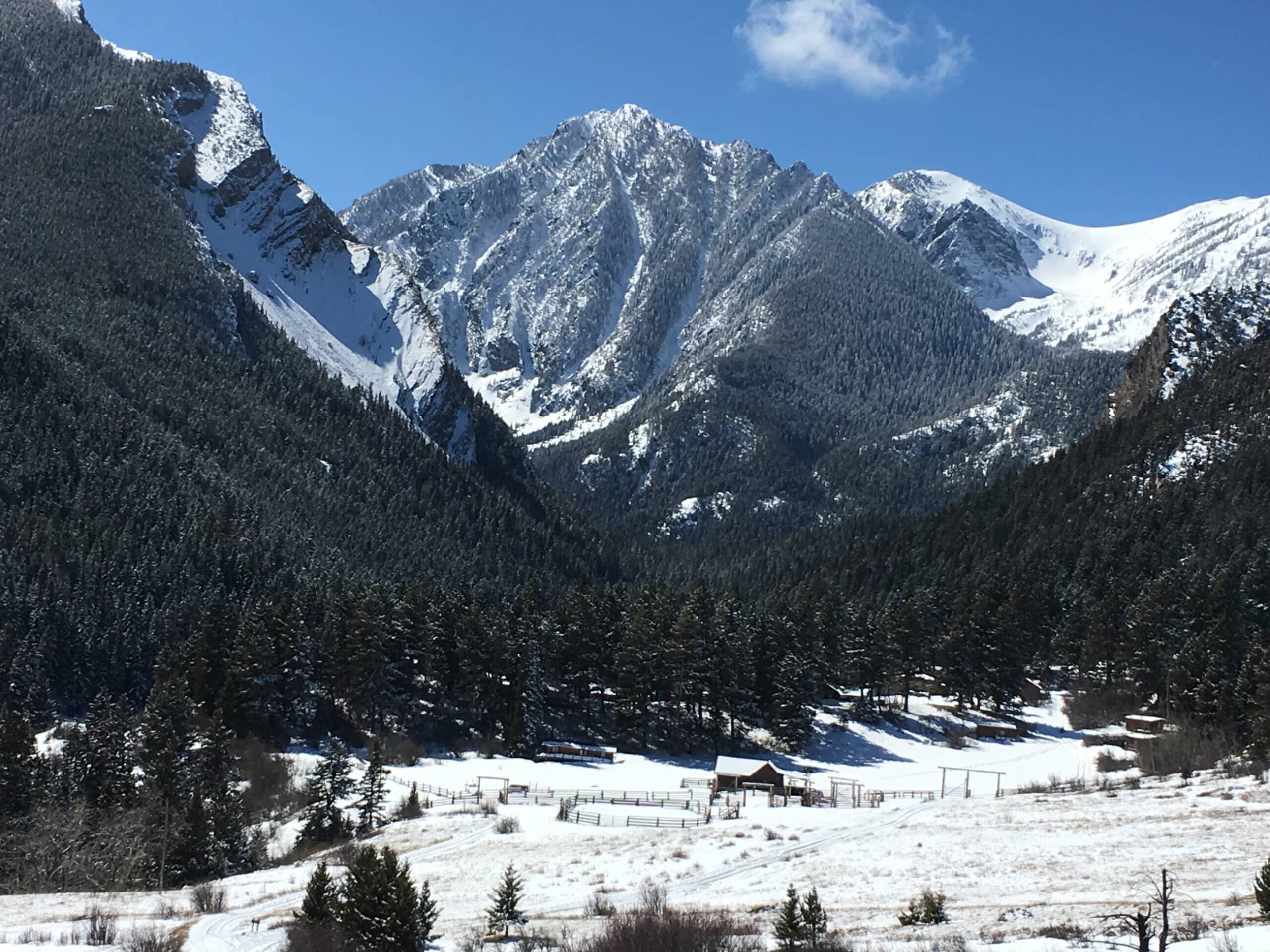 Snow-covered mountains with rugged peaks rise behind a forest of evergreens and a small building in a snowy valley under a clear blue sky, ideal for recreational land or hunting property.