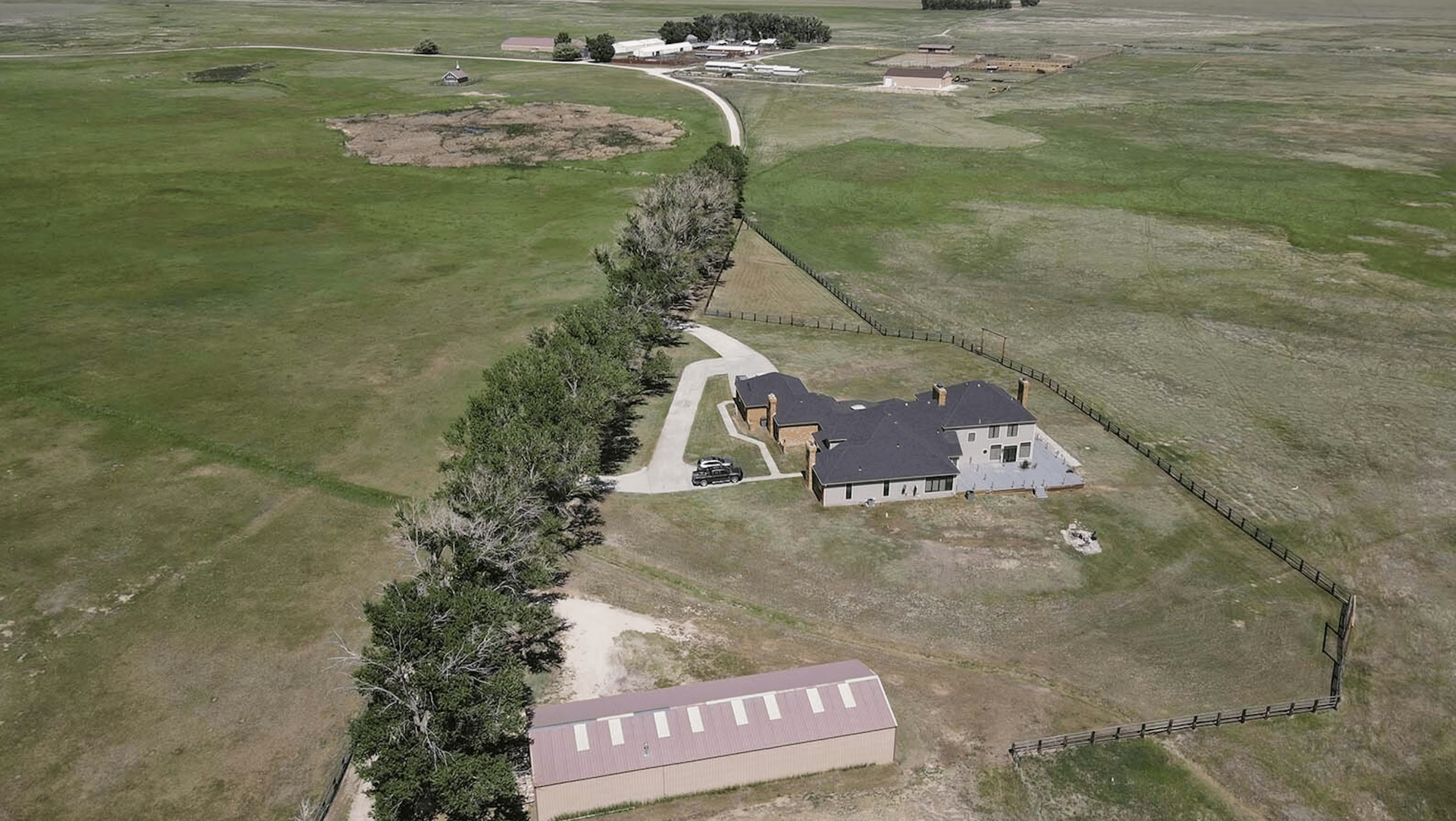 Aerial view of a rural cattle ranch with a main house, a driveway lined with trees, an outbuilding, and sprawling green fields. Additional farm buildings are visible in the distance.