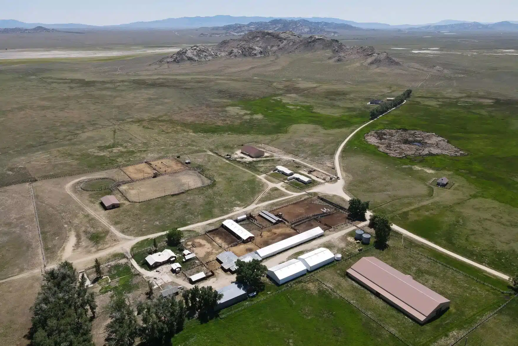 Aerial view of a cattle ranch with several buildings, barns, fenced enclosures, and green fields surrounded by open plains and rocky hills. Dirt roads connect the hunting property across the landscape.