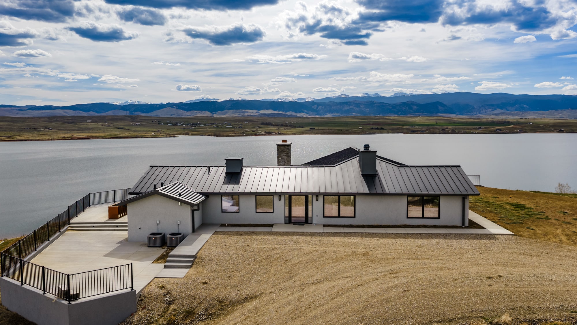 Modern single-story house with a metal roof overlooks a large lake and distant mountains under a partly cloudy sky; a concrete patio and railing border this beautiful ranch for sale.
