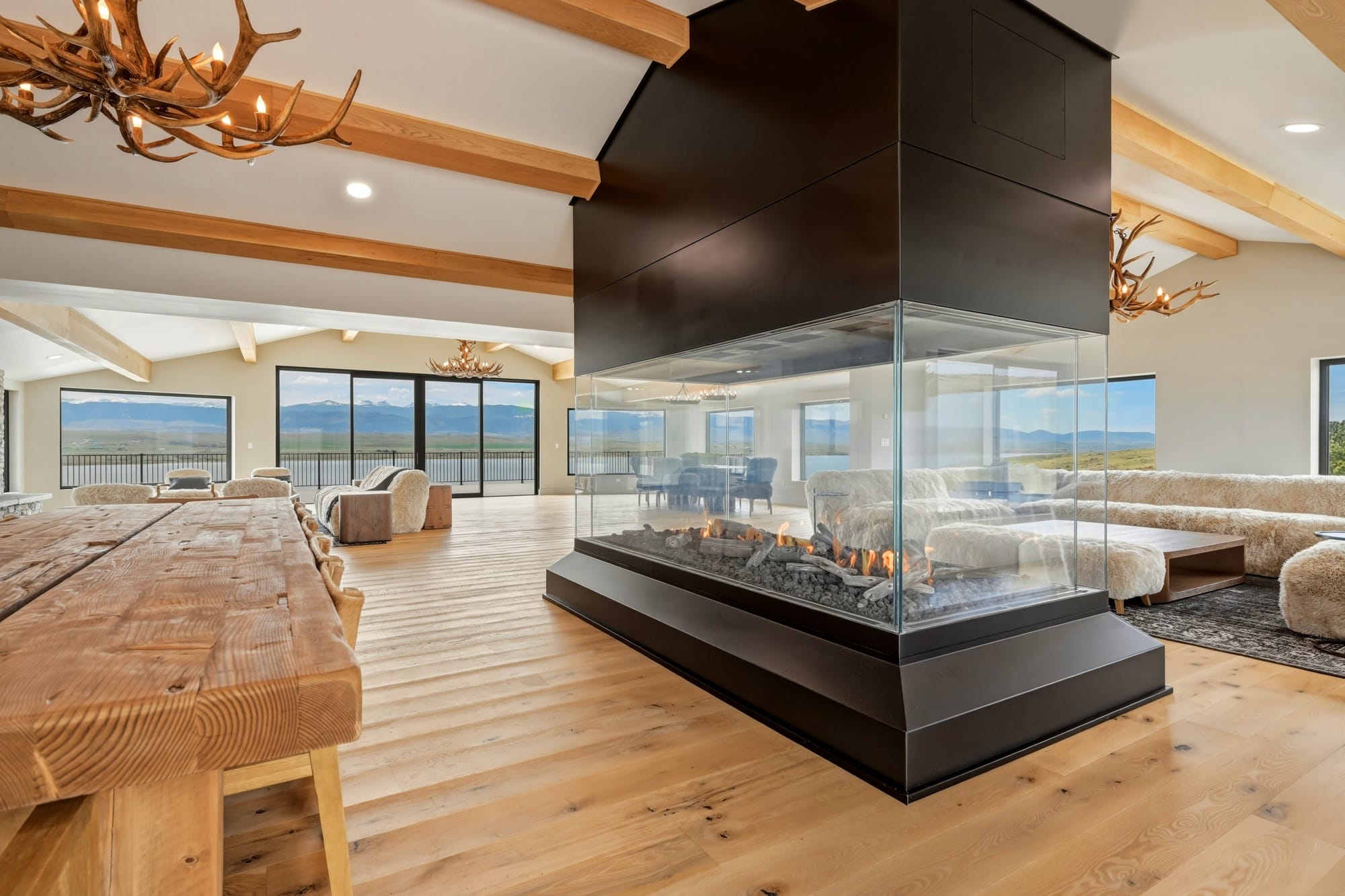Spacious modern living room with large windows, mountain views, wood floors, a central glass-enclosed fireplace, rustic wooden dining table, and antler chandeliers—perfect for your ranch for sale or recreational land retreat.
