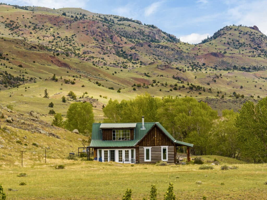 The Tom Miner Creek Retreat is a rustic wooden cabin with a green roof, nestled in a grassy field and surrounded by trees, rolling hills, and patches of shrubs with red and tan earth under a partly cloudy sky.