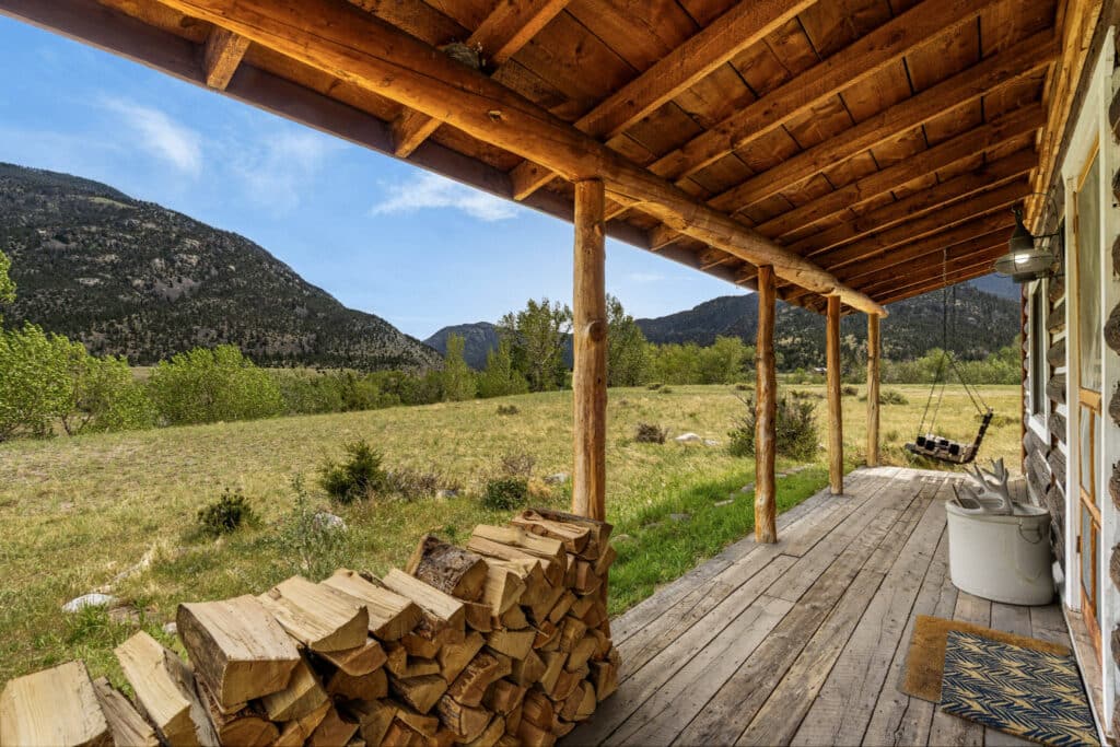 Wooden porch at this Montana retreat with a stack of firewood overlooks a grassy meadow and distant tree-covered mountains under a blue sky; a swing and rustic decor add to the cozy, peaceful Tom Miner Creek setting.