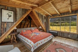 A cozy rustic bedroom with wood-paneled walls and ceiling, a large bed with a colorful quilt, side tables, and a large window offering a scenic Montana retreat view of mountains and fields. A painting hangs on the wall above the bed.