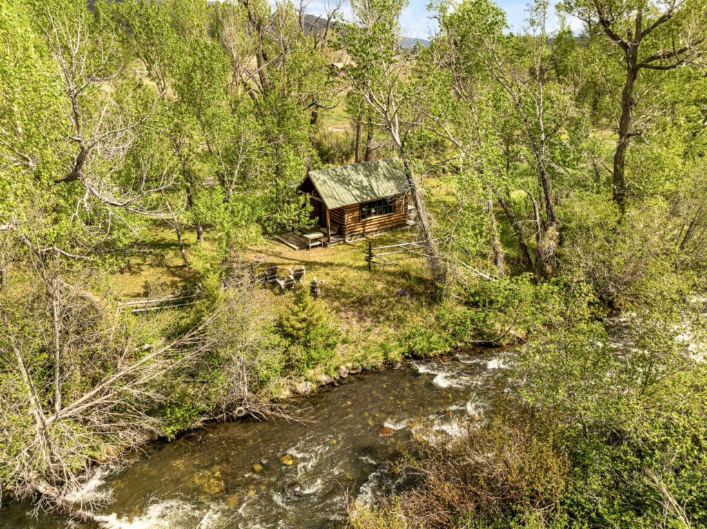 A small wooden cabin sits among tall green trees beside Montana’s Tom Miner Creek. Outdoor chairs and a table are arranged near the cabin, creating a peaceful retreat surrounded by lush vegetation under a bright, clear sky.