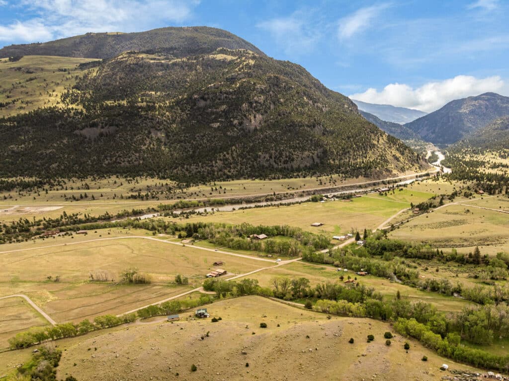 A wide landscape view of Montana’s Tom Miner Creek valley, scattered houses, green fields, and trees nestled among rolling hills and mountains—an inviting retreat beneath a partly cloudy blue sky.