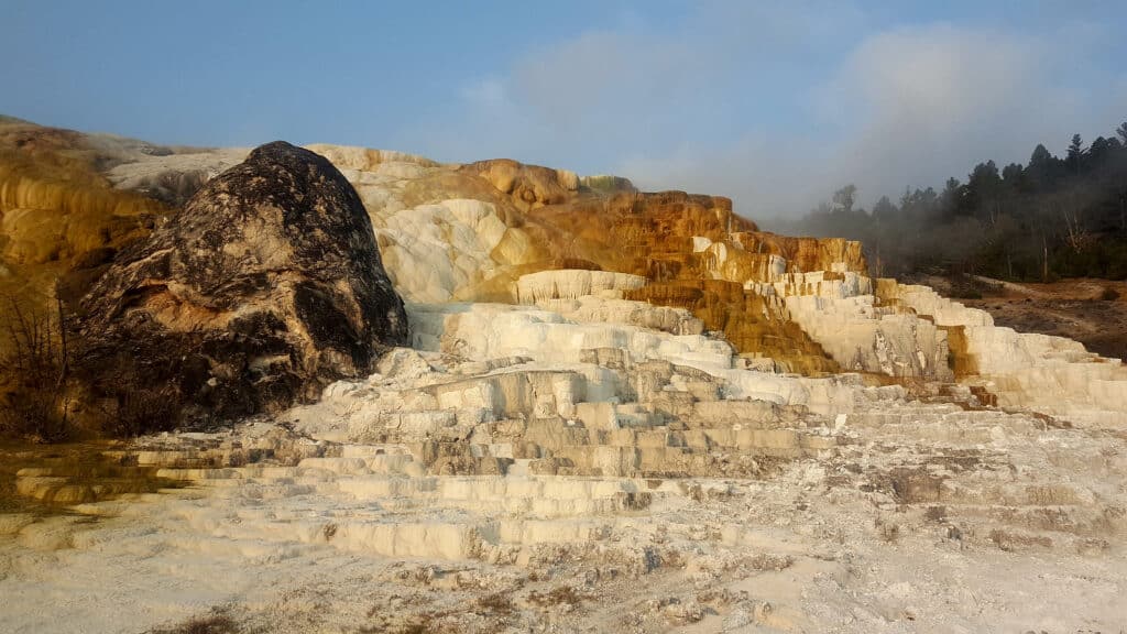 Terraced white and yellow mineral formations with a large dark rock, surrounded by sparse vegetation and trees under a blue sky with some clouds near Tom Miner Creek Retreat.