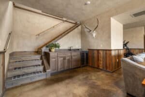 The basement at 380 Mill Gulch Road features rustic wood stairs, a built-in kitchenette with cabinets and mini fridge, a potted plant on the counter, and a mounted deer skull with antlers—plus glimpses of a sofa and exercise equipment.