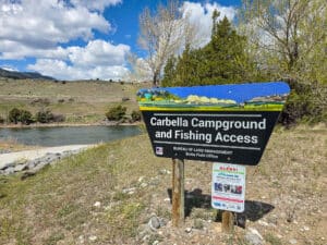 A sign for Carbella Campground and Fishing Access stands near a river in Montana, with grassy hills and trees in the background under a partly cloudy sky—a perfect retreat near Tom Miner Creek.