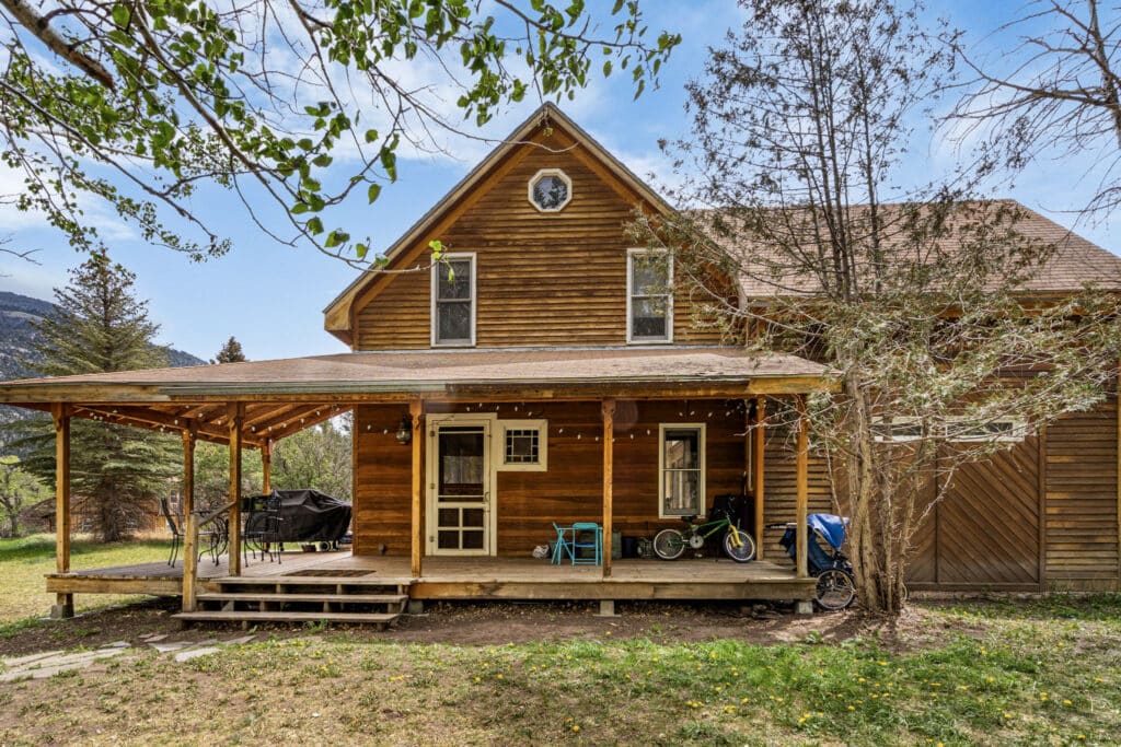 A rustic wooden house with a covered front porch, outdoor furniture, bikes, and a grill. Trees surround the yard at this Montana retreat near Tom Miner Creek, with mountains visible in the background under a blue sky.