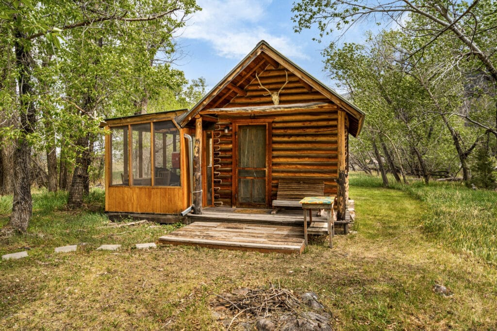 A small rustic log cabin retreat with a screened porch sits among green trees and grass under a blue sky near Tom Miner Creek, Montana, with a wooden bench and stone path leading to the front steps.