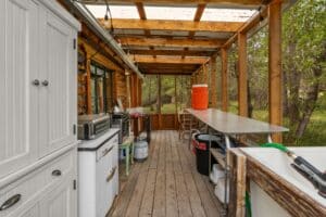 A rustic outdoor kitchen at a Montana retreat features wooden beams, a metal countertop, sink, appliances, storage shelves, and a bright orange drink cooler, overlooking green trees and grass by Tom Miner Creek. Natural light streams through a clear roof.