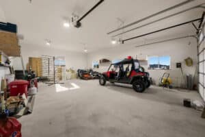 Spacious garage at 380 Mill Gulch with high ceilings, concrete floor, and overhead lights. Features shelves, tools, lawn equipment, a red utility vehicle, riding mowers, and supplies neatly organized along the walls. Sunlight enters through windows.