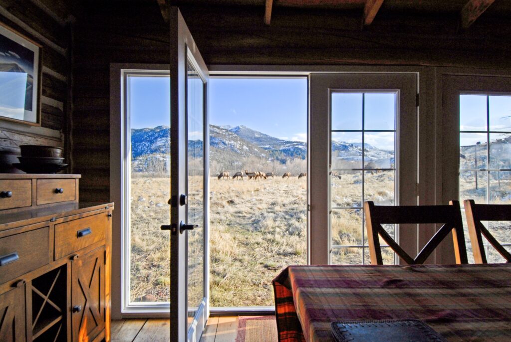 View from inside a rustic Montana cabin looking out glass doors onto a grassy field with a herd of elk and snow-capped mountains beyond. A dining table and chairs are in the foreground—perfect for a peaceful Tom Miner Creek retreat.