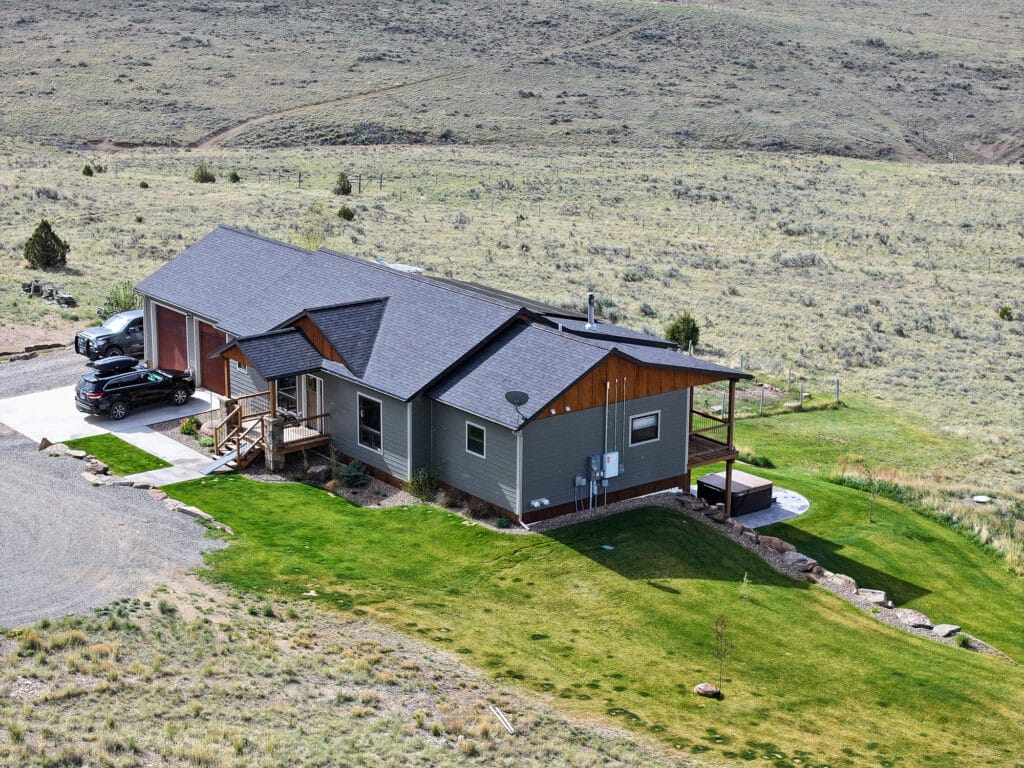 A modern single-story house with gray siding and a dark roof sits at 380 Mill Gulch Road in a rural grassy landscape. Several cars are parked beside it, and there is a covered porch and a small lawn surrounding the home.
