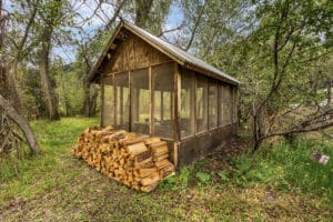 A rustic wooden cabin at Tom Miner Creek Retreat with screened windows sits in a green, wooded area. A neatly stacked pile of firewood rests beside the cabin, surrounded by grass, trees, and leafy vegetation.
