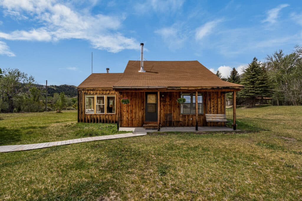 A rustic wooden cabin at Tom Miner Creek Retreat sits on a grassy lawn, surrounded by trees and hills under a partly cloudy blue sky. A wooden walkway leads you to the inviting front door and covered porch.
