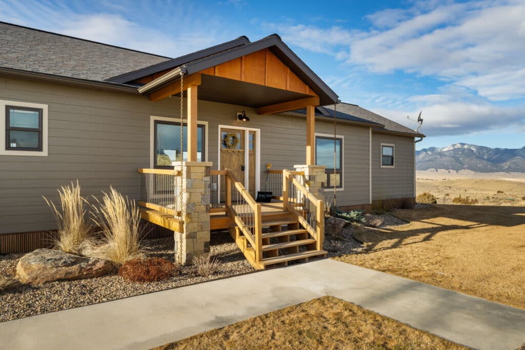 A modern single-story house at 380 Mill Gulch Road features a wooden front porch and steps, surrounded by dry grass and landscaping, set against a backdrop of mountains under a partly cloudy sky.