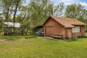 A wooden garage with a brown roof stands on a grassy lawn, surrounded by trees—a peaceful retreat near Tom Miner Creek, Montana. Next to it is a small shed with a metal roof and a carport for storing lawn equipment.