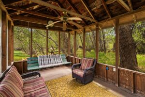 A cozy screened-in porch at this Montana retreat features wooden beams, two cushioned chairs, a green bench swing, a yellow patterned rug, and a ceiling fan—all surrounded by lush trees near scenic Tom Miner Creek.
