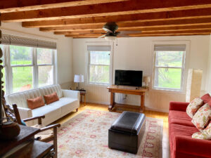 A cozy living room at Tom Miner Creek Retreat features wooden ceiling beams, a white couch, patterned rug, small TV on a wooden table, and large windows overlooking a green yard. Light fills the space, highlighting comfortable seating and simple decor.