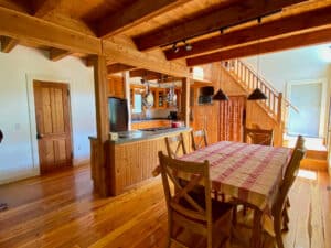 A cozy Montana kitchen and dining area with wooden floors, walls, and ceiling. A table with a striped cloth and six chairs sits in the foreground, while a staircase leads upstairs. Natural light fills this inviting retreat near Tom Miner Creek.
