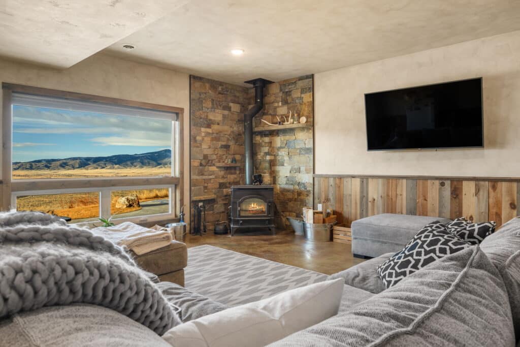Cozy living room at 380 Mill Gulch Road with a gray sectional sofa, patterned rug, wood-burning stove, wall-mounted TV, and large window offering a scenic view of mountains and fields. Rustic decor with stone and wood accents.