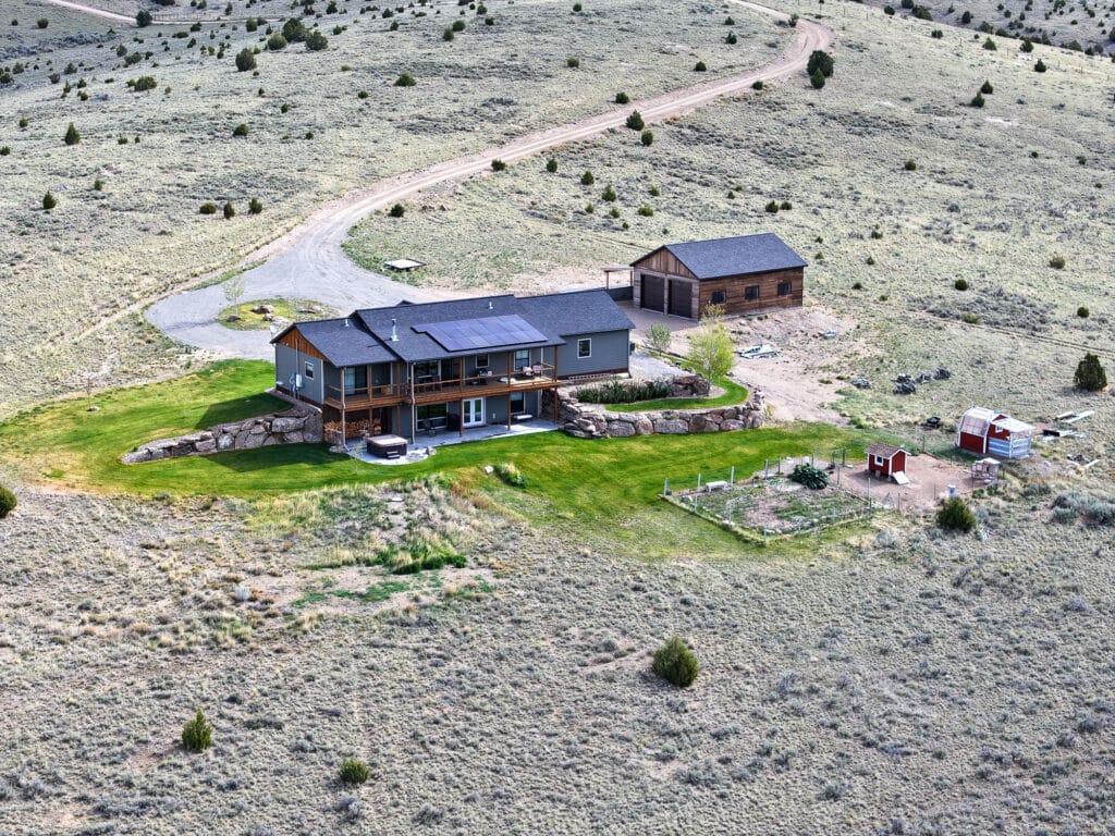 Aerial view of a modern house at 380 Mill Gulch Road with solar panels, a detached garage, a small fenced garden, and a red animal enclosure set in a dry, grassy landscape with sparse trees and winding dirt roads.